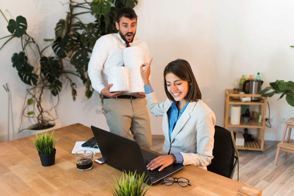 Young woman at laptop holds up hand to male co-worker with more work