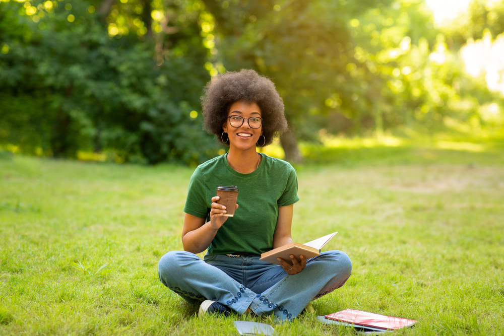 Woman taking a break outside with coffee, sitting on the grass with a book