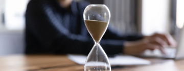 Image of an hourglass on a desk with a person in the background working