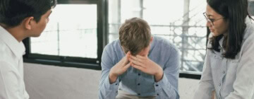 Man sitting with head in hands while two colleagues talk with him, demonstrating conflict resolution skills in a workplace discussion.