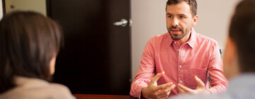 A young man facing the camera interviews for a sales representative job in a call center with two company representatives whose backs are to the camera, best jobs in Tijuana.