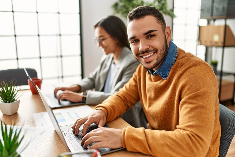 Smiling Latino African man working on a laptop with female coworker in background, best jobs in Tijuana.