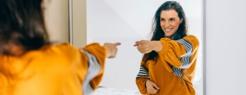 Employee smiling at their reflection in a mirror to reinforce positive self talk at work and build confidence.