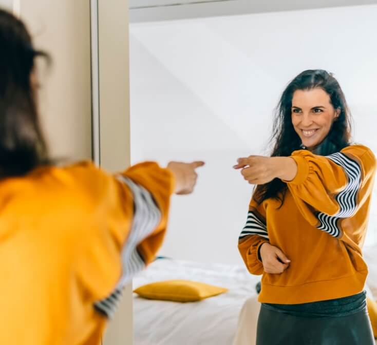 Employee smiling at their reflection in a mirror to reinforce positive self talk at work and build confidence.