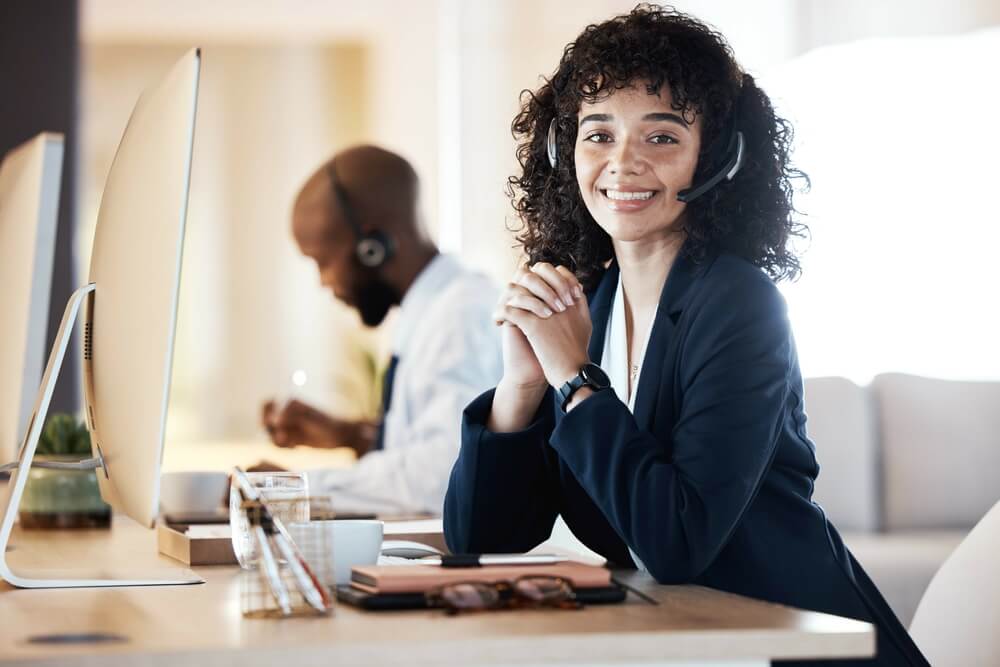 Young woman in call center with headset smiles at the camera while in the background a young man with a headset takes notes. Best call center jobs in Tijuana.