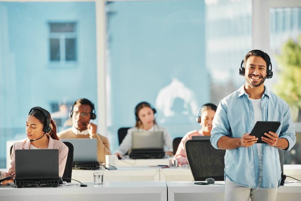 Young male supervisor smiles at camera while call center employees work behind him at laptops - best call center jobs in Tijuana.