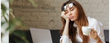 Sleepy worker with cup of coffee in her hand sitting in front of laptop - best call center jobs in Tijuana.