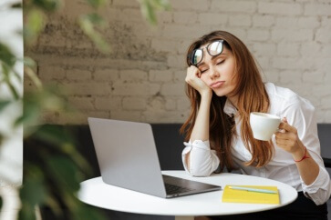 Sleepy worker with cup of coffee in her hand sitting in front of laptop - best call center jobs in Tijuana.