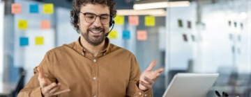 A smiling male call center worker speaks to a customer over the headset while multitasking tasks.
