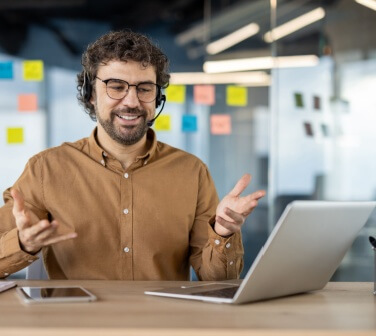 A smiling male call center worker speaks to a customer over the headset while multitasking tasks.
