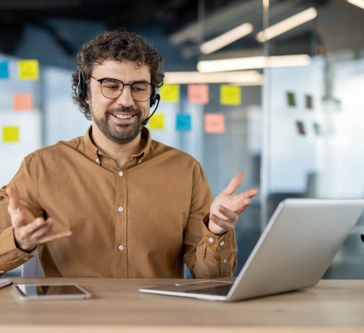 A smiling male call center worker speaks to a customer over the headset while multitasking tasks.
