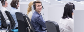 Male call center agent sitting in row of agents working in cubicles turns and smiles at camera - best call center jobs in Tijuana.