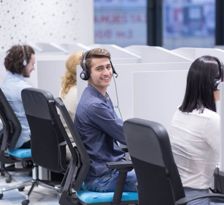 Male call center agent sitting in row of agents working in cubicles turns and smiles at camera - best call center jobs in Tijuana.