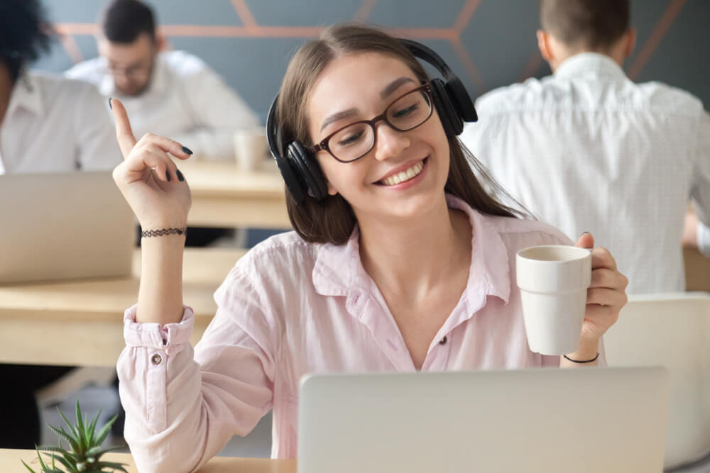 Happy millennial woman in headphones at call center and drinking coffee, smiling female call center agent enjoying working with customers.