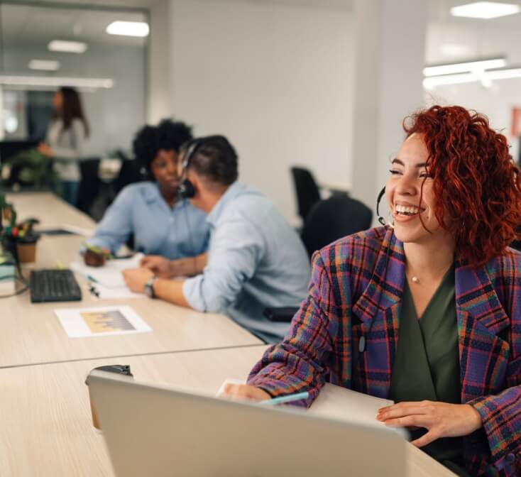Smiling customer services agent woman with headset working in an office. Portrait of a female agent working on a laptop in a call center and talking with clients and customers.