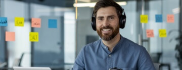 Portrait of a smiling young male call center worker in headphones working in the office at a laptop, holding a cup of coffee and looking at the camera, confidence building at work.