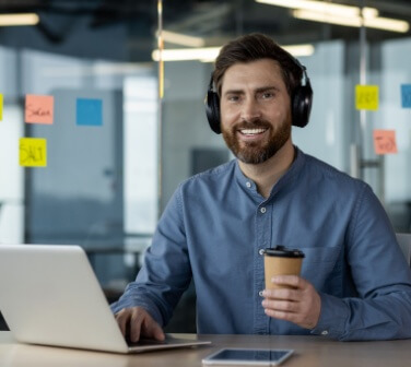 Portrait of a smiling young male call center worker in headphones working in the office at a laptop, holding a cup of coffee and looking at the camera, confidence building at work.