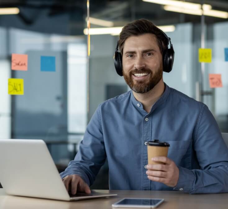 Portrait of a smiling young male call center worker in headphones working in the office at a laptop, holding a cup of coffee and looking at the camera, confidence building at work.