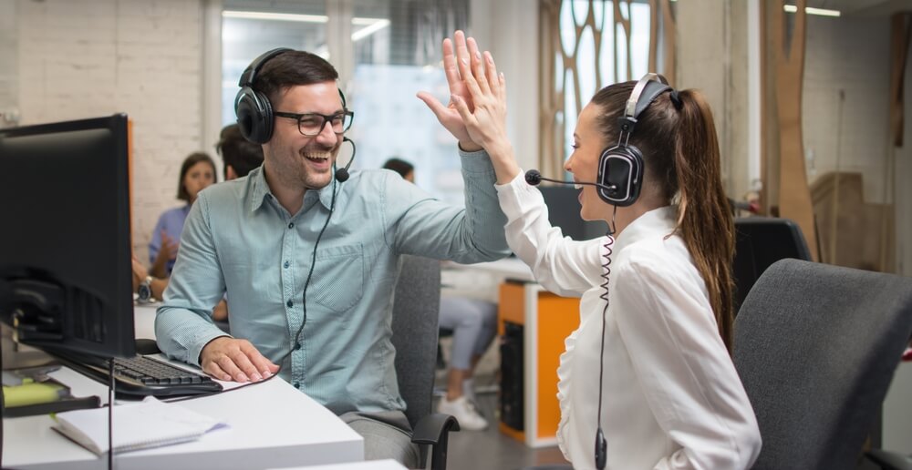 Two smiling agents at their work station, a woman and a man, with problem solving skills in call centers celebrate with a high five. 