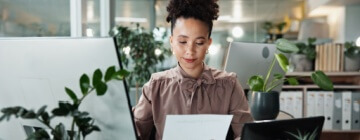 Business, woman and paperwork by computer in office for data entry in CRM system and reading revenue report. Account representative, employee and document for client sale stats and portfolio review.