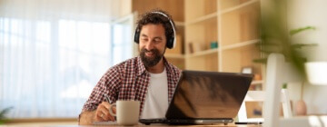 Cheerful man sitting at a wooden table with a laptop and headphones, enjoying a cup of coffee while working taking advantage of remote career opportunities in Tijuana.