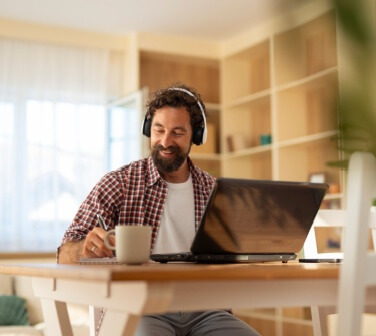Cheerful man sitting at a wooden table with a laptop and headphones, enjoying a cup of coffee while working taking advantage of remote career opportunities in Tijuana.