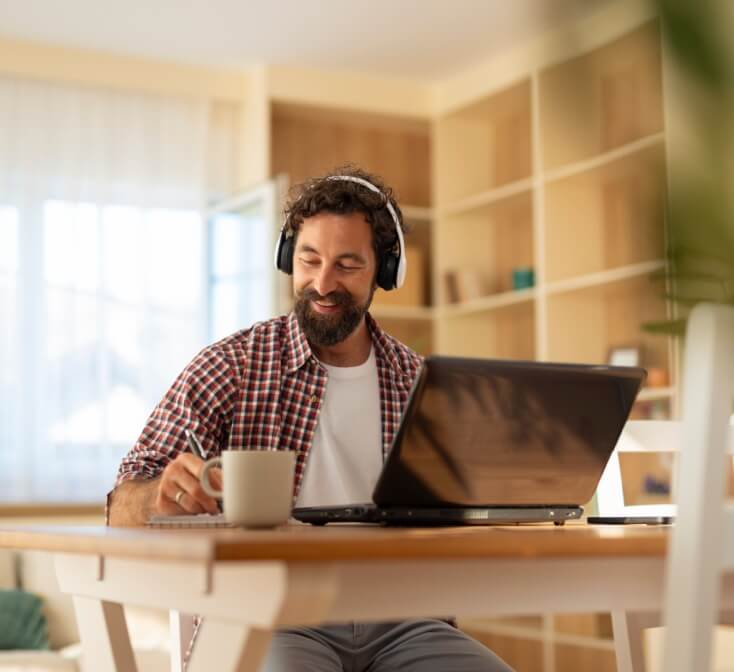 Cheerful man sitting at a wooden table with a laptop and headphones, enjoying a cup of coffee while working taking advantage of remote career opportunities in Tijuana.