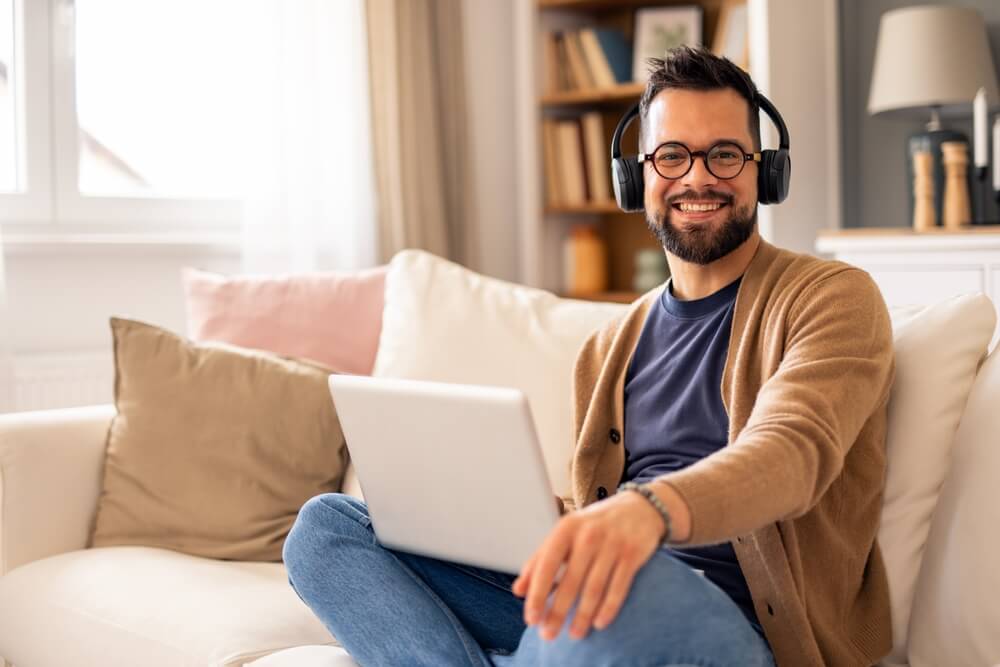 A cheerful man sits on a couch with headphones, working on a laptop in a bright, cozy living room with soft lighting as part of remote career opportunities in Tijuana.
