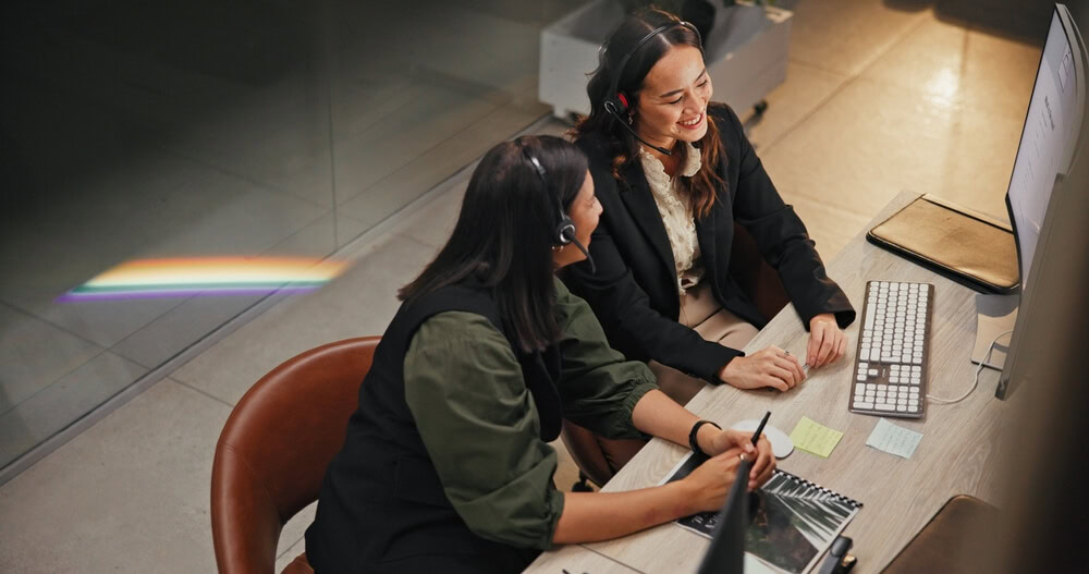 Two call center professionals wearing headsets collaborate at a desk while reviewing notes and a computer screen, illustrating teamwork and growth within call center career paths.