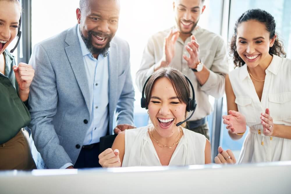 A call center agent wearing a headset celebrates at her desk while teammates applaud behind her, highlighting success and support in the call center first 90 days.