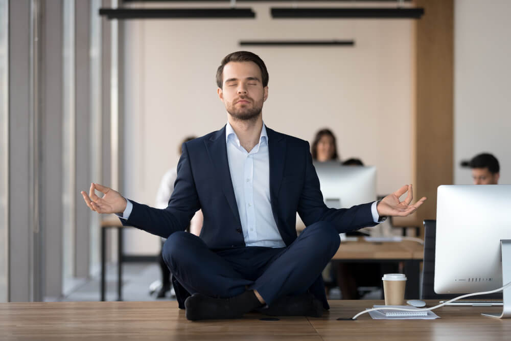 Business professional meditating at desk in open office, illustrating calm focus and conflict resolution skills in a workplace environment.