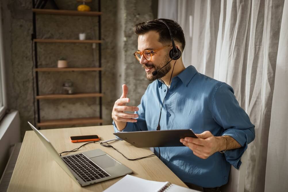 Man wearing glasses and a headset speaks during a video call while holding a tablet at a home office desk, reflecting call center recruiter expectations.