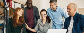 Diverse group of coworkers smiles and collaborates around a laptop at a meeting table, reflecting teamwork while working with U.S. clients.