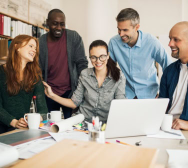 Diverse group of coworkers smiles and collaborates around a laptop at a meeting table, reflecting teamwork while working with U.S. clients.