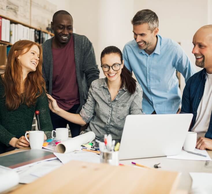 Diverse group of coworkers smiles and collaborates around a laptop at a meeting table, reflecting teamwork while working with U.S. clients.