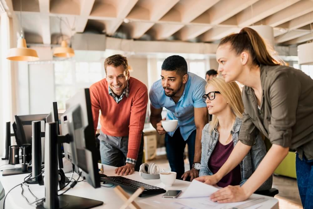 Team of coworkers gathers around multiple computer monitors in a modern office, reviewing documents and on-screen data while working with U.S. clients.