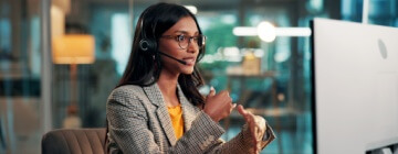 Call center agent demonstrating call center communication skills while speaking on a headset and gesturing beside a desktop computer in a modern office.
