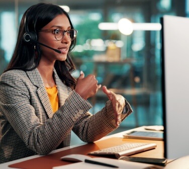 Call center agent demonstrating call center communication skills while speaking on a headset and gesturing beside a desktop computer in a modern office.