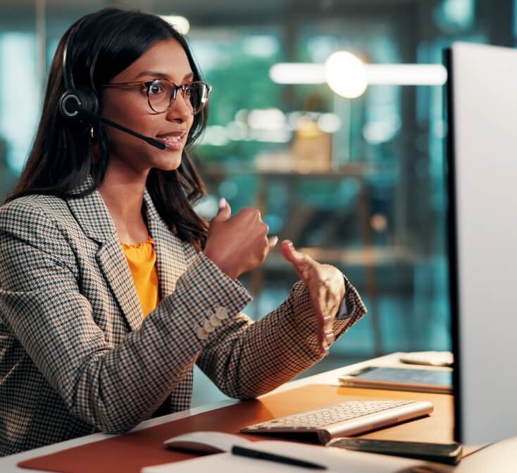 Call center agent demonstrating call center communication skills while speaking on a headset and gesturing beside a desktop computer in a modern office.