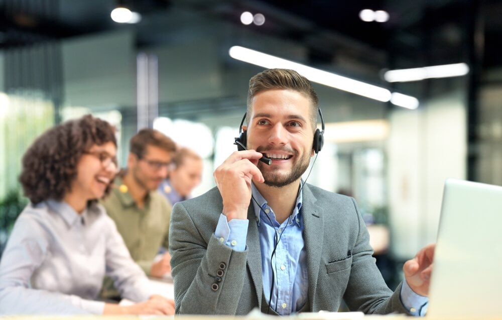 Call center agent smiling while wearing a headset at his desk, highlighting call center communication skills in a modern office setting.