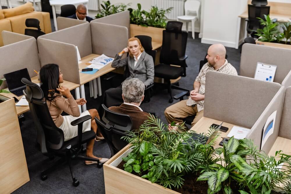 Employees gathered in a modern office workspace with cubicles and indoor plants, creating a collaborative and homey office environment.
