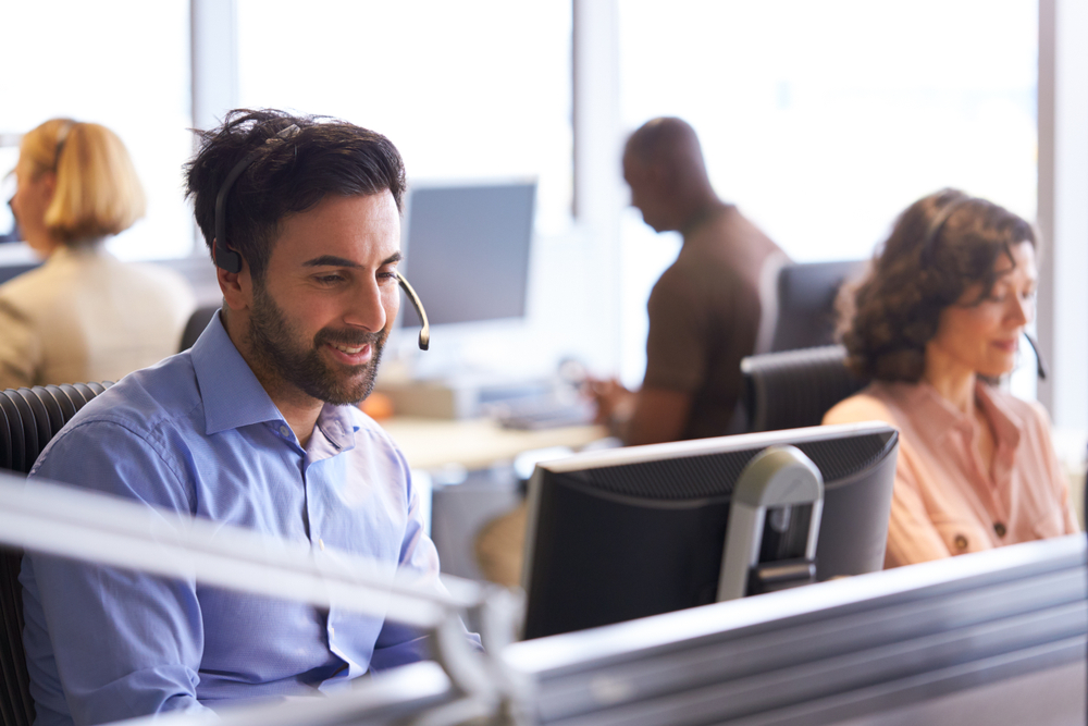 Businessman Wearing Telephone Headset Talking To Caller In Multicultural Call Center Customer Services Department.