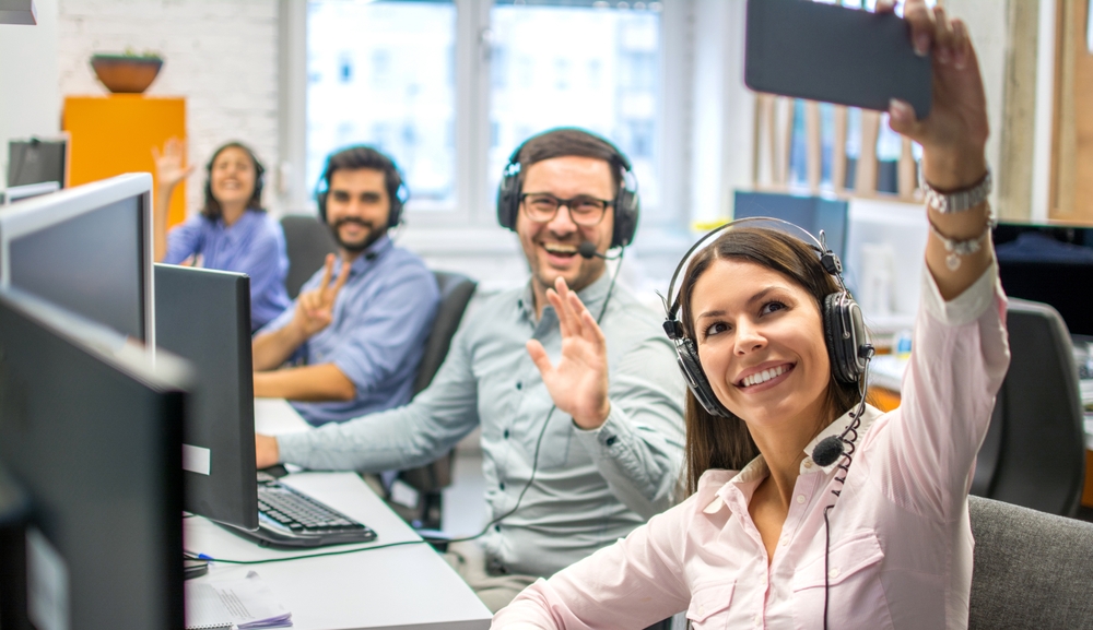Woman customer support operator taking a selfie photo of her team colleagues in a multicultural call center. 