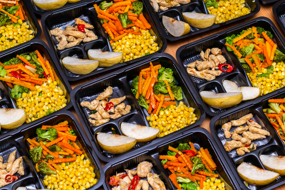 Meal prep containers filled with chicken, vegetables, corn, and sliced fruit.