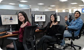 Three employees sit at computer workstations in an office and wave at the camera.