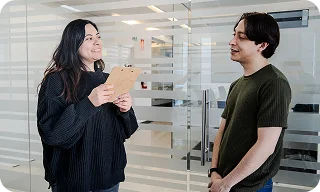 Two coworkers stand in an office, talking while one holds a clipboard.