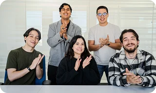 A group of coworkers smiles and claps together while sitting and standing around a table in an office.