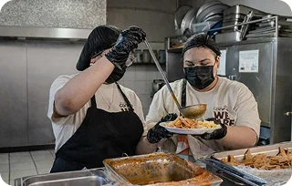 Two workers wearing gloves and face masks serve food from trays in a kitchen.