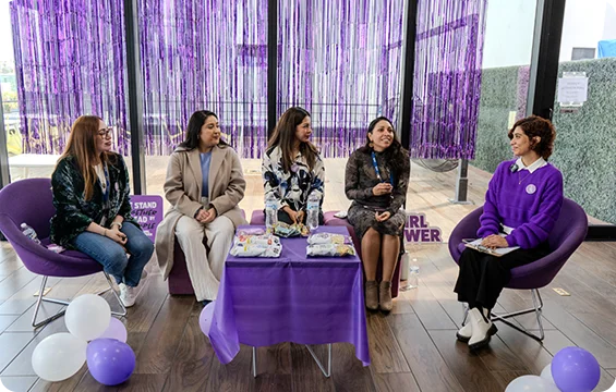A group of women sit together on chairs during a panel discussion in a decorated event space with purple accents.